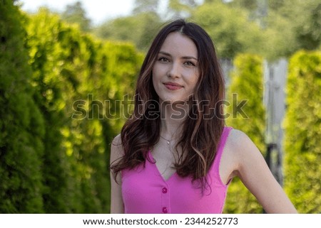 Portrait of a beautiful young woman model with wavy brunette hair in a pink dress against a background of green bushes
