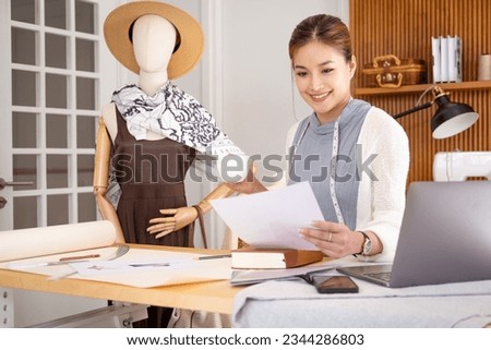 Happy female stylist or tailor work with paperwork use laptop at workplace. Smiling young asian businesswoman fashion designer sit at modern office desk consider document drink coffee in morning.