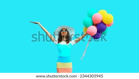 Summer image of happy smiling young woman with bunch of colorful balloons having fun wearing straw hat, dress on blue studio background