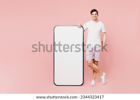 Full body young smiling fun caucasian man wear white t-shirt casual clothes big huge blank screen mobile cell phone smartphone with area looking camera isolated on plain pastel light pink background