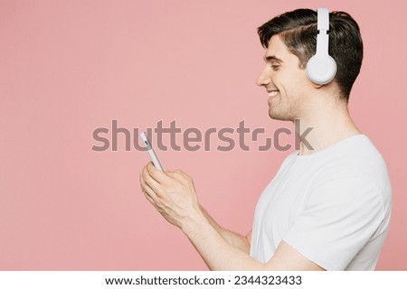 Side view young happy caucasian man wear white t-shirt casual clothes listen to music in headphones look aside on area isolated on plain pastel light pink background studio portrait. Lifestyle concept