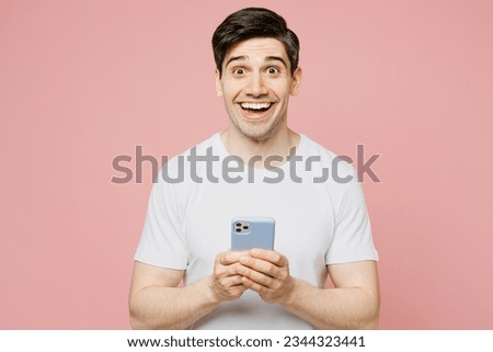 Young surprised caucasian man wearing white t-shirt casual clothes hold in hand using mobile cell phone in blue case isolated on plain pastel light pink background studio portrait. Lifestyle concept