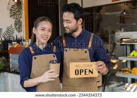 Small business couple husband and wife startup lifestyle concept, Asian man and woman wearing apron standing in front of counter holding coffee cup and open sign, coffee shop owner partnership
