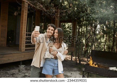 Smiling couple with wine taking selfie on smartphone near bbq and vacation house at background