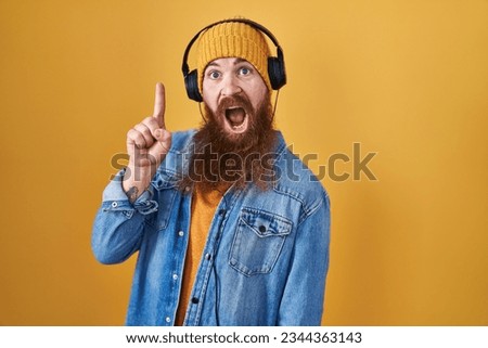 Caucasian man with long beard listening to music using headphones pointing finger up with successful idea. exited and happy. number one. 