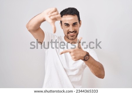Handsome hispanic man standing over white background smiling making frame with hands and fingers with happy face. creativity and photography concept. 