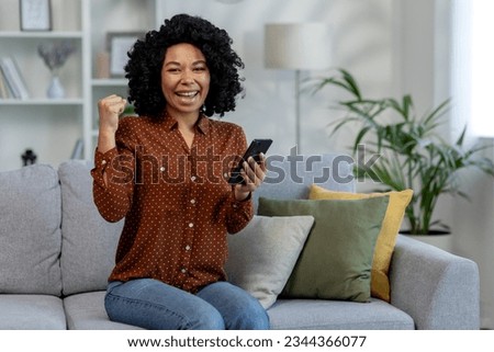 Happy African American woman holding phone while sitting on sofa at home and looking happy at camera showing victory gesture with hand.