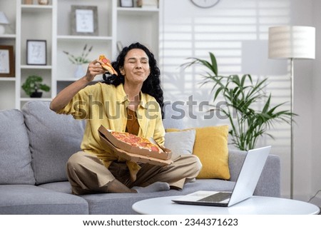 Joyful and happy woman eating pizza alone at home, Hispanic woman satisfied with timely food delivery, using laptop to watch online video stream, sitting on sofa in living room at home.