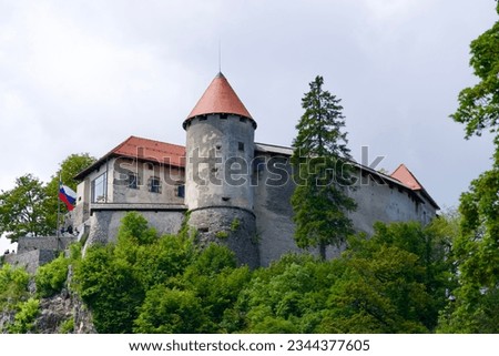 Castle on top of rock with waving Slovenian flag at lakeshore of Lake Bled on a cloudy summer day. Photo taken August 8th, 2023, Bled, Slovenia.
