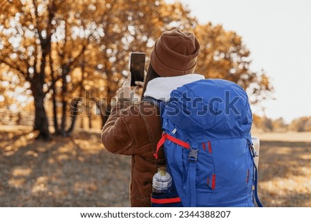 Female backpacker with smartphone taking photo in autumn forest during travel 