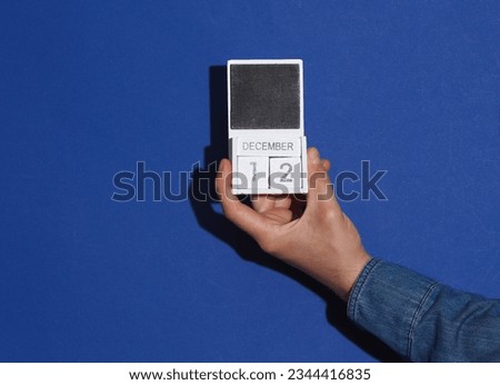 Man's hand in denim shirt holds wooden block calendar organizer with date December 12 on a blue background