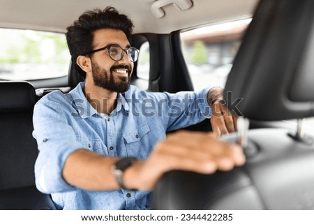 Happy handsome bearded young indian guy in casual outwear sitting on backseat inside car taxi, looking through window and smiling, going to office or to airport. Eastern man travelling by auto Royalty-Free Stock Photo #2344422285