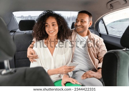 Joyful Cab Experience. Smiling Arab Passengers Couple Enjoying Comfortable Car Ride In Taxi, Lady Holds Smartphone with Transfer Transportation Application, Spouses Hugging On Backseat In Auto Royalty-Free Stock Photo #2344422331