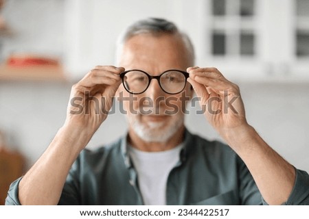 Eye Vision Concept. Smiling Senior Man Looking At Camera Through Eyeglasses In His Hands, Handsome Elderly Gentleman Trying New Glasses While Standing At Home Interior, Selective Focus