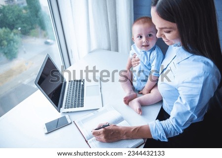 High angle view of beautiful business lady holding her cute little baby, making notes and smiling while working in office. Baby is looking on his mom