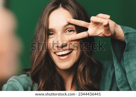 Beautiful young brunette curly woman taking selfie, posing with peace v-sign, smiling happy, take photo, posing against green background.