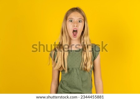 Oh my God. Surprised Caucasian kid girl wearing green T-shirt over yellow background stares at camera with shocked expression exclaims with unexpectedness,