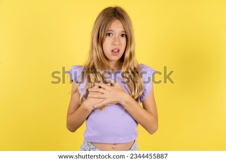 Scared Caucasian kid girl wearing violet T-shirt over yellow background looks with frightened expression, keeps hands on chest, being puzzled to notice something strange, People, hush reaction and emo
