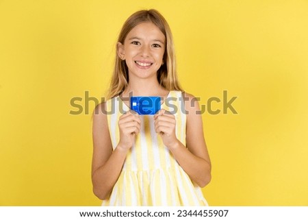 Photo of cheerful Caucasian kid girl wearing yellow dress over yellow background hold debit card look empty space