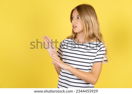Displeased Caucasian kid girl wearing striped T-shirt over yellow background keeps hands towards empty space and asks not come closer sees something unpleasant