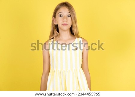 Stunned Caucasian kid girl wearing yellow dress over yellow background stares reacts on shocking news. Astonished preteen girl holds breath Royalty-Free Stock Photo #2344455965