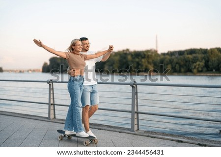 Joyful couple smiling and looking away while driving skateboard close to river bridge. Their hands are widely opened.