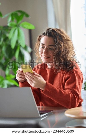 Young happy pretty woman sitting at table holding smartphone using cellphone modern technology, looking at mobile phone while working or learning, texting messages, browsing web at home. Vertical