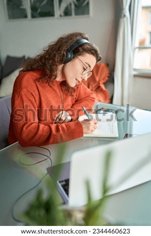 Smiling young woman university student wearing headphones using laptop elearning, writing notes, studying online education class by webinar, learning online seminar sitting at home table. Vertical
