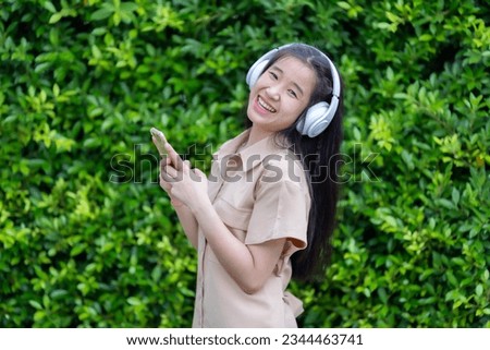 young asian girl using smartphone holding mobile phone device with wearing white wireless headphones and listening to music and using in the park outdoors or at the cafe in the sunny weather day