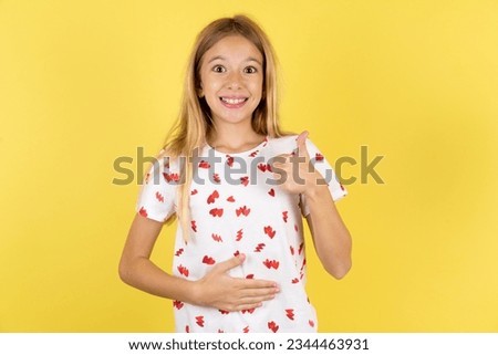 Caucasian kid girl wearing polka dot shirt over yellow background happy positive smile hands on belly show thumb-up fine healthy