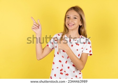 Caucasian kid girl wearing polka dot shirt over yellow background indicating finger empty space showing best low prices, looking at the camera