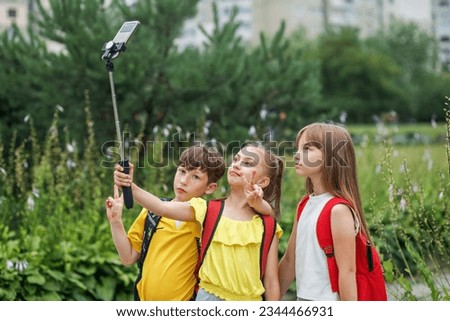 Group of elementary school students with backpacks taking picture by smartphone on selfie stick outdoor. Primary education