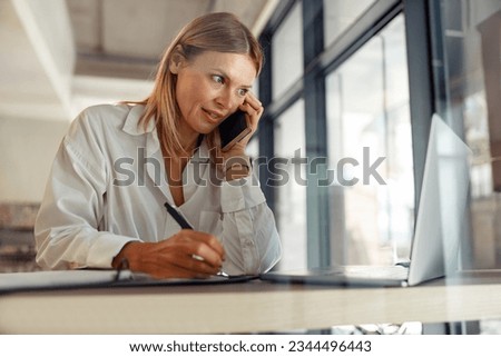 Woman entrepreneur making notes and talking by phone with client while sitting in coworking  Royalty-Free Stock Photo #2344496443