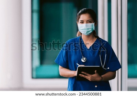 Asian female nurse or doctor wearing blue scrubs and face mask holding a clipboard in hospital. Royalty-Free Stock Photo #2344500881