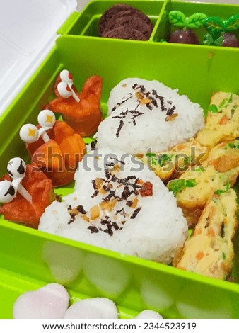 Photo of a school lunch menu, consisting of heart-shaped rice, fried sausages, omelet, crackers, biscuits and grapes
