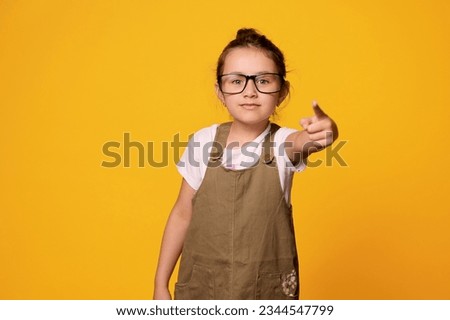 Serious primary school student girl, first grader wearing teacher's black framed glasses, points her index finger at camera, showing warning hand sign, isolated orange background. Back to school