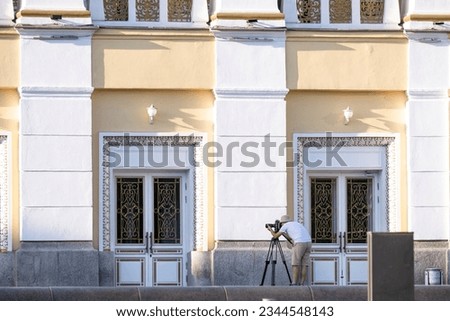 The photographer looks into the eyepiece of a camera mounted on a tripod against the backdrop of an old building.