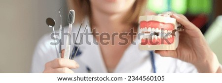 Female stomatologist shows dentist tools and toothbrush in one hand and jaws with teeth in another. Dental clinic and healthcare concept. Royalty-Free Stock Photo #2344560105