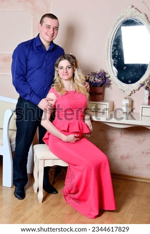 Attractive young couple is celebrating holiday at home. Standing and sitting on the background of a mirror. Husband hugs his pregnant wife. Family standing and sitting near a table with a mirror. High