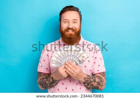 Photo of positive cheerful man with redhair beard wear stylish pink t-shirt hold bunch of dollars isolated on blue color background