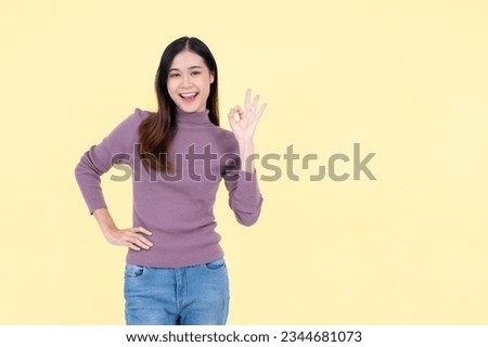 A portrait of a beautiful and smiling young Asian woman in casual clothes shows the Okay hand sign and stands against an isolated yellow background. Agree, deal, confirm, perfect