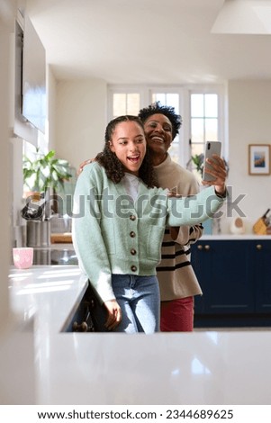 Mother With Teenage Daughter At Home In Kitchen Posing For Selfie On Mobile Phone