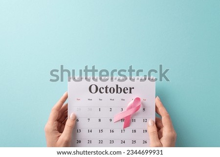 Health in focus: First person top view of female hands holding October calendar, prioritizing doctor's appointment for breast cancer screening. Pink ribbon on pastel blue background with space for ad Royalty-Free Stock Photo #2344699931