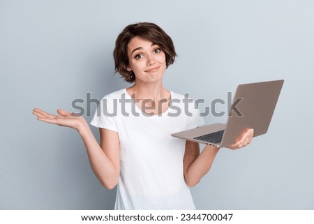 Photo of clueless girl with bob hairstyle dressed white t-shirt holding laptop shrugging shoulders isolated on gray color background