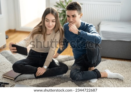 Teenage couple taking a selfie photo while sitting together on the floor at home.