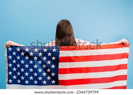 Young smiling happy woman wears casual clothes holding american flag and looking to the camera isolated on blue color background