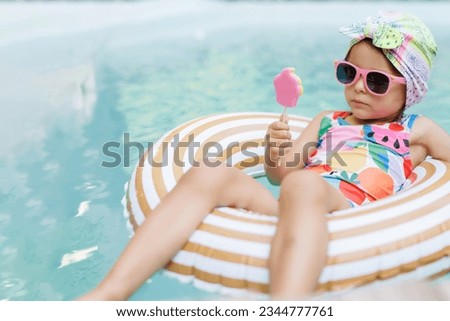 Cute funny toddler girl in colorful swimsuit and sunglasses relaxing on inflatable toy ring floating in pool have fun during summer vacation in tropical resort. Child having fun in swimming pool. 