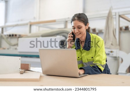 Female worker using laptop computer and talking with client by smartphone at furniture factory.