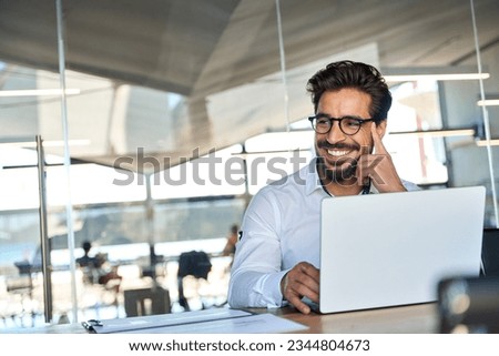 Happy Latin business man wearing glasses working at laptop in office looking away. Happy young male professional using computer sitting at desk thinking of corporate technology solutions. Copy space Royalty-Free Stock Photo #2344804673