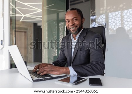 Portrait of successful happy african american boss, man smiling and looking at camera, businessman in business suit sitting at desk with laptop inside office. Royalty-Free Stock Photo #2344826061